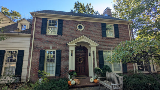 A front-facing view of a brick house with dark shutters, a wooden front door, a bench in the yard, and pumpkins near the entrance.