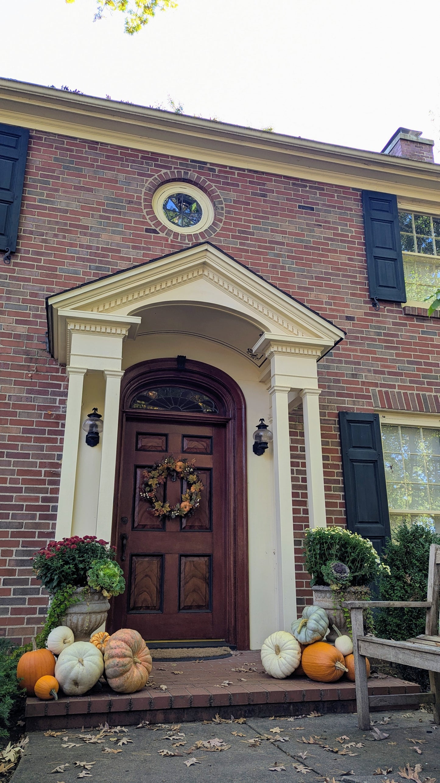 A front porch of a brick house decorated for autumn, with a dark wooden door, white columns, pumpkins, potted plants, and a wreath on the door.