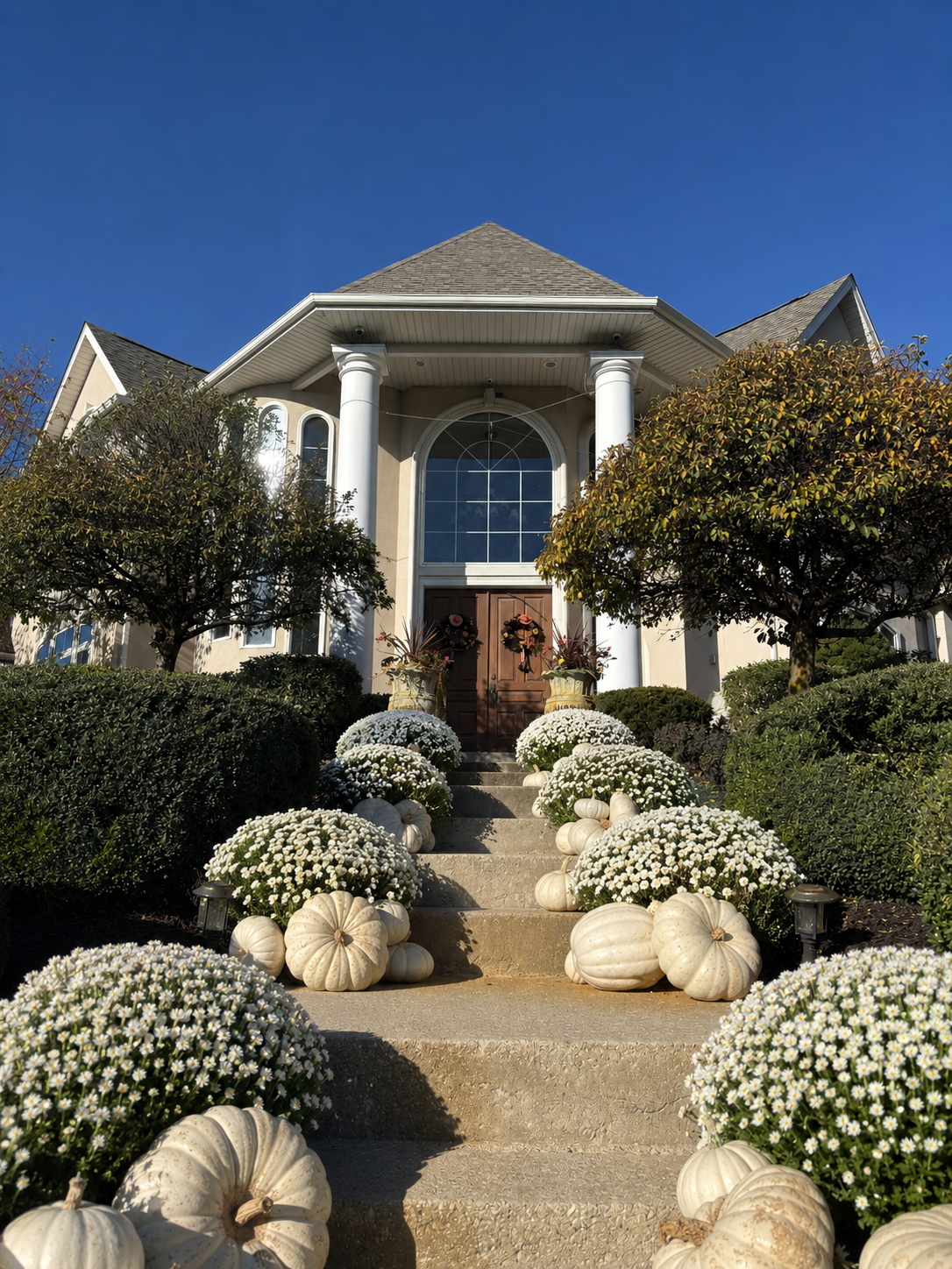 A front yard staircase leading to a house entrance, decorated with white mums, pumpkins, and potted plants under a clear blue sky.